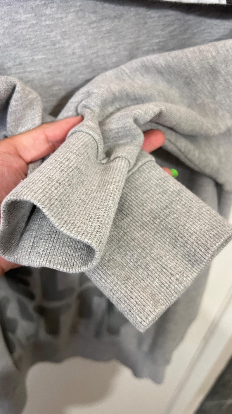 May include: Close-up of a gray sweatshirt sleeve. The ribbed cuff is visible, along with the soft, textured fabric of the sleeve. The sweatshirt appears to be made of a comfortable material, suitable for casual wear.