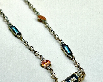 Puede incluir: Collar plateado con una serie de detalles rectangulares y ovalados de esmalte en tonos azules, naranjas y rojos. El collar tiene un colgante cilíndrico con una base de esmalte azul oscuro y un diseño floral. La cadena está hecha de pequeños eslabones interconectados.
