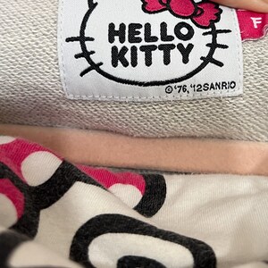 May include: Close-up of a gray sweatshirt label featuring the Hello Kitty logo in black and a pink bow. The text "HELLO KITTY" is prominently displayed. The label also includes the copyright information.