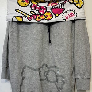 May include: Gray hooded sweatshirt dress featuring a large Hello Kitty graphic on the front. The top portion has a white collar with a colorful pattern of bows, donuts, and the words "Hello Kitty". The dress has long sleeves and a drawstring.