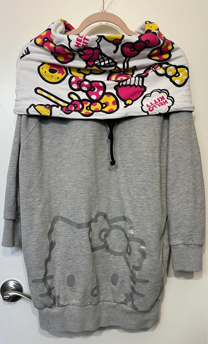 May include: Gray hooded sweatshirt dress featuring a large Hello Kitty graphic on the front. The top portion has a white collar with a colorful pattern of bows, donuts, and the words "Hello Kitty". The dress has long sleeves and a drawstring.