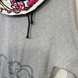 May include: Gray sweatshirt featuring a Hello Kitty graphic on the front. The cowl neck is white with a Hello Kitty print in pink, yellow, and black. The sweatshirt has black drawstrings.
