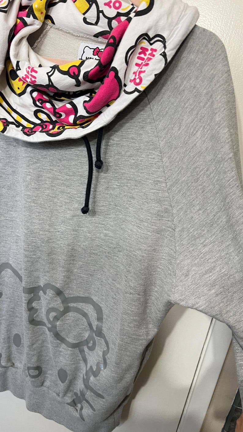 May include: Gray sweatshirt featuring a Hello Kitty graphic on the front. The cowl neck is white with a Hello Kitty print in pink, yellow, and black. The sweatshirt has black drawstrings.