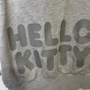 May include: A gray sweatshirt with the words "HELLO KITTY" in large, gray, block letters across the front. The sweatshirt has a ribbed hem and cuffs.