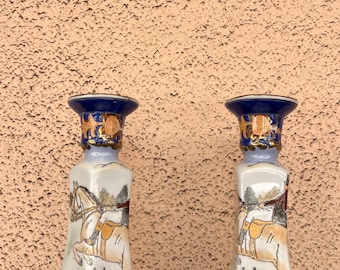 May include: Pair of decorative ceramic candle holders. Each holder features a blue and gold rim, a painted scene of a rider on a horse, and a black and white dog. The holders are approximately 10 inches tall.