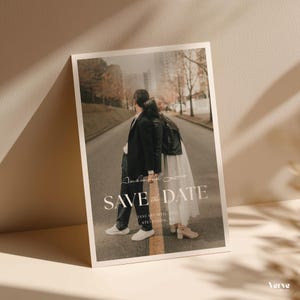 Pode incluir: Um cartão Save the Date com uma fotografia de um casal numa estrada. O cartão apresenta as palavras "Save the Date" em uma fonte elegante. A data e o local também estão incluídos.