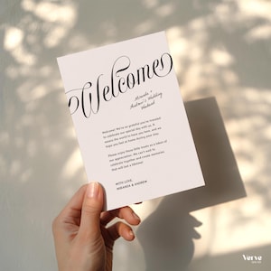 May include: A white wedding welcome card held by a hand. The card features the word "Welcome" in elegant script, along with the names "Miranda & Andrew" and the text "Wedding Weekend". The card includes a message of gratitude and appreciation.