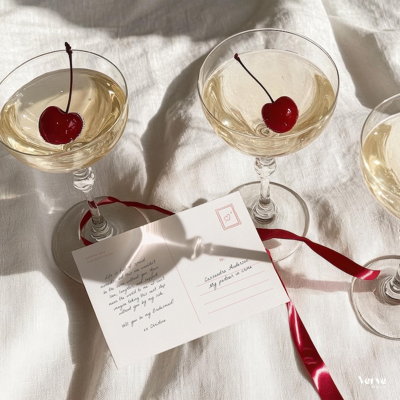 May include: Champagne glasses with cherries, a postcard, and a red ribbon on a white surface. The postcard has text that reads "Will you be my Bridesmaid?" and "My partner in crime."