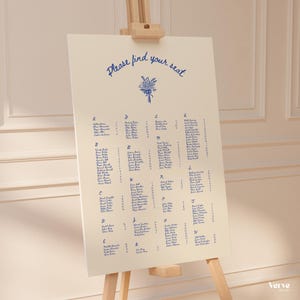 Puede incluir: Un plano de asientos crema con texto de caligrafía azul sobre un caballete. El texto dice "Please find your seat" encima de una ilustración floral. Los nombres están listados alfabéticamente por apellido. El caballete es de madera clara.