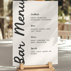 Può includere: Un menu da bar rettangolare bianco con testo nero che recita Bar Menu, Cocktails, Beer, Wine e Spirits, elencando varie opzioni di bevande. Il menu è tenuto in posizione verticale da un supporto in legno chiaro su una superficie piana con ombre.