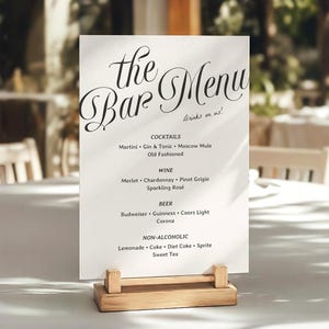 Op de afbeelding: Een wit rechthoekig barmenu-bord met zwarte tekst die the Bar Menu, Drinks on us! leest, inclusief lijsten met cocktails, wijn, bier en alcoholvrije dranken. Het bord wordt rechtop gehouden door een lichtgekleurde houten blokstandaard.