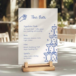 May include: A white rectangular bar menu card in a light wood stand featuring blue line art of a hand holding a glass and a champagne tower. The text reads The Bar, Signature Drink, The Sweet Start, Beer, Budweiser, Guinness, Coors Light, Corona, Wine, Merlot, Chardonnay, Pinot Grigio, Sparkling Rose, Spirits, Vodka, Scotch, Whiskey, Gin, Bourbon.