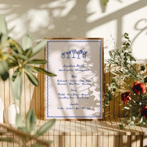 A bar menu card with a blue border and text, stands on a wooden shelf surrounded by plants and floral arrangements at a wedding reception. The menu lists signature drinks, beer, wine, and spirits.