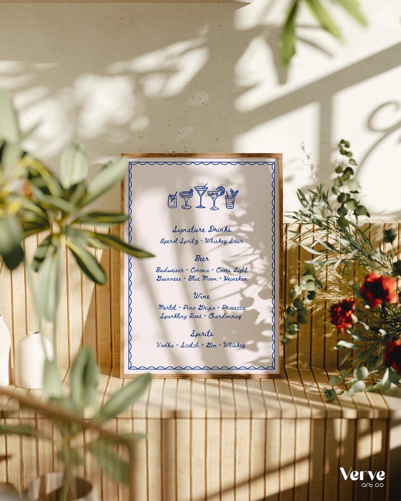 A bar menu card with a blue border and text, stands on a wooden shelf surrounded by plants and floral arrangements at a wedding reception. The menu lists signature drinks, beer, wine, and spirits.