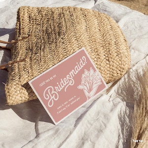 May include: A woven straw handbag rests on a white surface next to a pink invitation card. The card reads "Will you be my Bridesmaid?" in elegant script, with floral illustrations and event details.