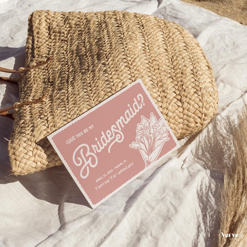 May include: A woven straw handbag rests on a white surface next to a pink invitation card. The card reads "Will you be my Bridesmaid?" in elegant script, with floral illustrations and event details.