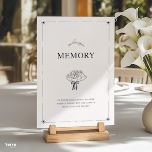 May include: A white memorial sign with black text, including the words "In Loving Memory" and a floral illustration. The sign is held by a light wood stand. The sign reads "Of those who cannot be here with us today, but are always with us in our hearts."