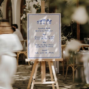 A wooden easel displays a bar menu with a blue hand drawn border, hand drawn drink illustrations, and handwritten text. The menu lists signature drinks, beer, wine, and spirits. The background shows an outdoor setting with blurred figures and greenery, suggesting a wedding or event.