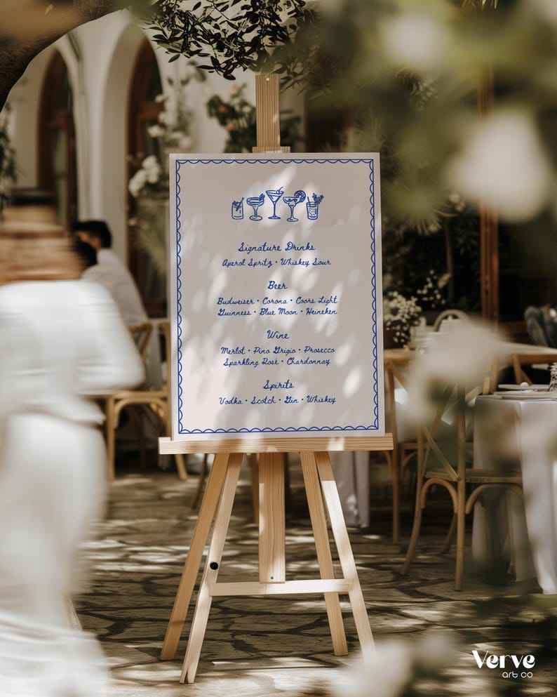 A wooden easel displays a bar menu with a blue hand drawn border, hand drawn drink illustrations, and handwritten text. The menu lists signature drinks, beer, wine, and spirits. The background shows an outdoor setting with blurred figures and greenery, suggesting a wedding or event.