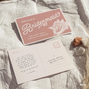 May include: Two pink postcards with the text "Will you be my Bridesmaid?" in a script font. One card has a floral illustration. The other card has handwritten text. The cards are on a white surface.