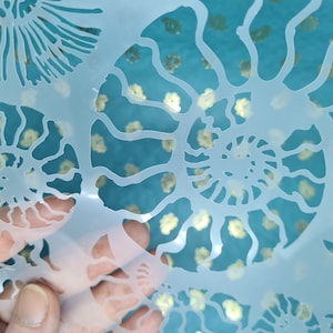 May include: A white plastic stencil featuring a design of multiple nautilus shells. The stencil is translucent, revealing a blue background with gold glitter.