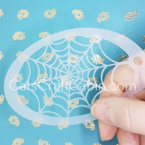 May include: A white plastic spiderweb stencil for crafting and decorating. The stencil is oval shaped and has a hole in the top for hanging.