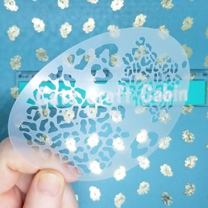 May include: A white plastic stencil with a leopard print design. The stencil is oval shaped and has a smooth surface.