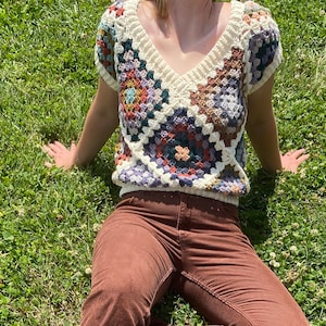 Puede incluir: Un top blanco de crochet con un patrón geométrico en tonos marrones, azules y verdes. El top tiene cuello en V y mangas cortas.