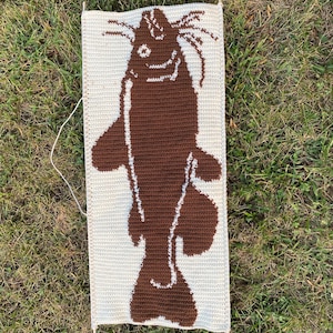 May include: A handmade, rectangular crochet wall hanging featuring a brown catfish design on a cream-colored background. The catfish is depicted in a simple, cartoonish style. The piece is held in place by wooden dowels at the top and bottom.