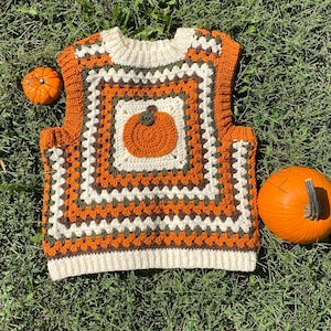 Könnte beinhalten: Handgestrickte Weste im Granny-Square-Muster in Orange-, Weiß- und Brauntönen. Auf der Vorderseite ist eine Kürbisapplikation zentriert. Zwei Kürbisse liegen neben der Weste auf dem Gras. Die Weste ist ein Kleidungsstück zum Thema Herbst.