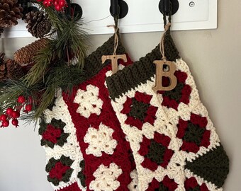 May include: Three handmade Christmas stockings hang from a white rack. The stockings are crocheted in a granny square pattern with red, cream, and green yarn. Wooden letters "T" and "B" are attached. A festive wreath with pinecones and berries is on the left.