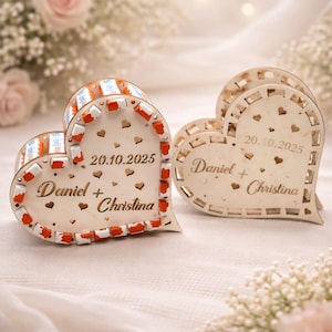 May include: Two heart-shaped wooden boxes with the names "Daniel + Christina" and the date "20.10.2025" engraved on them. One box has a decorative border with red and white accents. The boxes are on a white surface.
