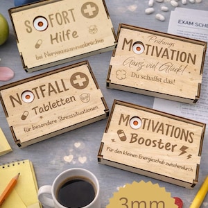 Puede incluir: Cuatro cajas de madera con texto y diseños grabados, incluyendo un símbolo de cruz e iconos de pastillas. Las cajas tienen frases como "SOFORT Hilfe" y "MOTIVATIONS Booster". También se ve una taza de café, un bloc de notas y pastillas.