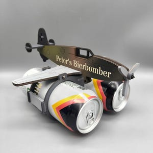 Könnte beinhalten: Ein dunkelbraunes Flugzeugmodell aus Holz, das dazu dient, zwei Getränkedosen zu halten. An der Seite des Rumpfes befindet sich der Schriftzug 'Peter's Bierbomber' in weißer Schrift sowie ein kleines Kreuzemblem am Heckbereich.