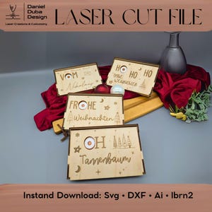 Pode incluir: Arquivo de corte a laser com quatro caixas de madeira com texto e desenhos festivos alemães. As caixas estão dispostas sobre uma tábua de madeira com tecido vermelho e ornamentos. O texto inclui "Vom Nikolaus", "Frohe Weihnachten", "Ho Ho Ho" e "Oh Tannenbaum".