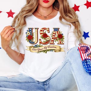 Może przedstawiać: Biała koszulka z napisem "USA" w kolorach czerwonym, białym i niebieskim, ozdobiona kwiatowymi akcentami. Tekst "250 Years Of Freedom" jest wyświetlany na banerze poniżej.