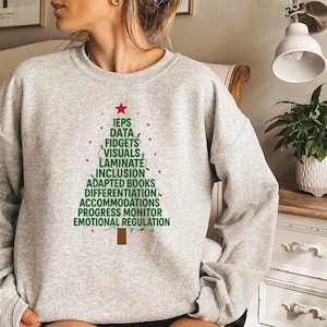 May include: A light gray crewneck sweatshirt featuring a graphic of a Christmas tree formed by stacked green text including IEPS, DATA, FIDGETS, VISUALS, LAMINATE, INCLUSION, ADAPTED BOOKS, DIFFERENTIATION, ACCOMMODATIONS, PROGRESS MONITOR, and EMOTIONAL REGULATION.