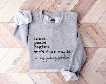 May include: A light gray crewneck sweatshirt featuring black text that reads: inner peace begins with four words: not my fucking problem. The garment has long sleeves with ribbed cuffs and a ribbed neckline, with a Gildan brand label visible inside.