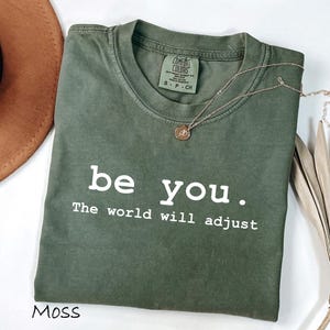 May include: A moss green short-sleeve t-shirt with a crew neckline and a white graphic print that reads "be you. The world will adjust". The shirt features a Comfort Colors brand label at the inner neck and is made of cotton fabric.