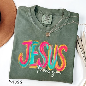 Puede incluir: Una camiseta verde musgo con las palabras "JESUS loves you" en un dise&ntilde;o colorido y en capas. La palabra "JESUS" est&aacute; en rosa brillante, azul y amarillo. Un collar dorado est&aacute; drapeado sobre la camisa.