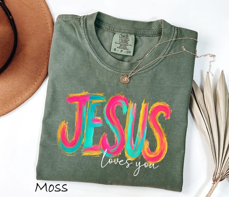 Puede incluir: Una camiseta verde musgo con las palabras "JESUS loves you" en un dise&ntilde;o colorido y en capas. La palabra "JESUS" est&aacute; en rosa brillante, azul y amarillo. Un collar dorado est&aacute; drapeado sobre la camisa.