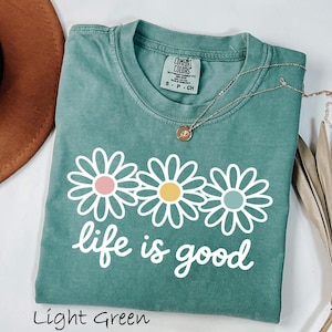May include: A light green short-sleeve t-shirt featuring a graphic of three white daisies with pink, yellow, and light green centers. The text "life is good" is printed in white below the flowers. The shirt has a Comfort Colors brand label.