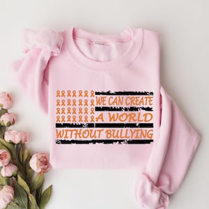 May include: A light pink crewneck sweatshirt featuring a graphic design of orange awareness ribbons arranged in a flag pattern. The text reads WE CAN CREATE A WORLD WITHOUT BULLYING in orange letters against black horizontal distressed stripes.