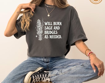Op de afbeelding: Een antracietgrijs T-shirt met korte mouwen, voorzien van een witte afbeelding van een saliebundel en de tekst "WILL BURN SAGE AND BRIDGES AS NEEDED." Het shirt heeft een ronde hals en een relaxte pasvorm, gecombineerd met een blauwe jeans en sneakers.