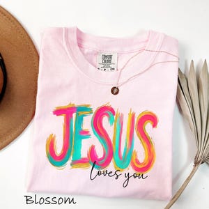 Puede incluir: Camiseta rosa claro con la frase "JESUS loves you" en un dise&ntilde;o colorido de pinceladas. La camiseta tiene cuello redondo y un peque&ntilde;o collar con colgante redondo. La palabra "Blossom" est&aacute; escrita en la parte inferior.
