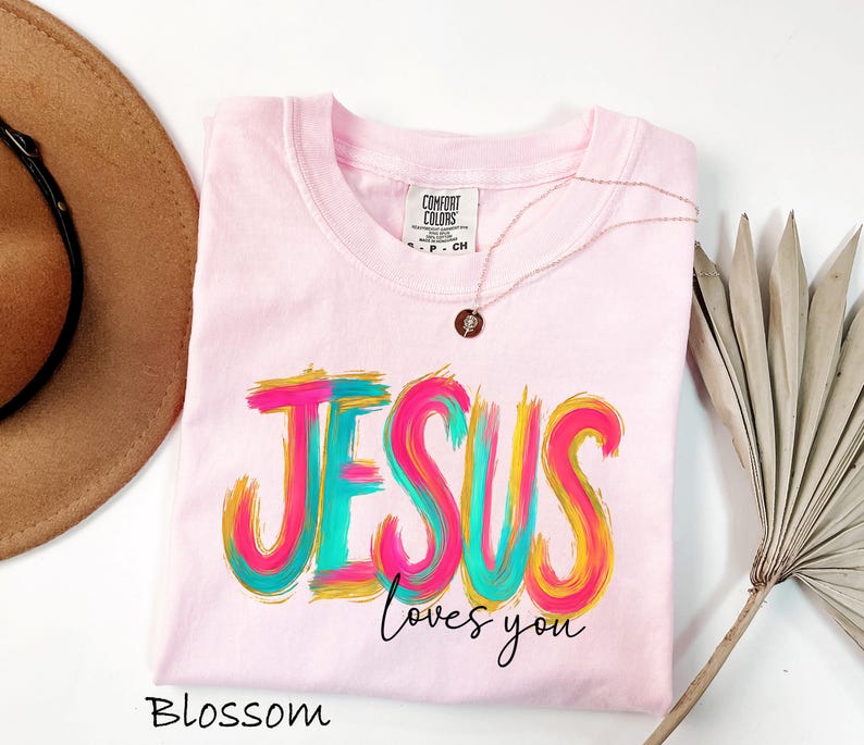Puede incluir: Camiseta rosa claro con la frase "JESUS loves you" en un dise&ntilde;o colorido de pinceladas. La camiseta tiene cuello redondo y un peque&ntilde;o collar con colgante redondo. La palabra "Blossom" est&aacute; escrita en la parte inferior.