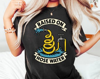 May include: A black t-shirt featuring a graphic of a yellow coiled garden hose connected to a metal spigot, with blue water spraying out. The text "RAISED ON" appears in a banner above the hose, and "HOSE WATER" is in a banner below the graphic.
