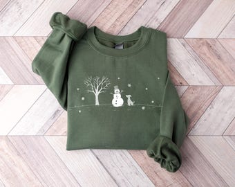 May include: An olive green crewneck sweatshirt featuring a white line art graphic of a winter scene. The design includes a bare tree, a snowman wearing a top hat, a small dog standing nearby, and scattered snowflakes on a horizontal ground line.
