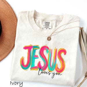 Puede incluir: Camiseta de color marfil con las palabras "JESUS loves you" en un dise&ntilde;o colorido de pinceladas. El texto es de color rosa brillante, turquesa y amarillo. Un collar de oro con un colgante descansa sobre la camiseta. Un sombrero marr&oacute;n y una planta seca est&aacute;n en el fondo.
