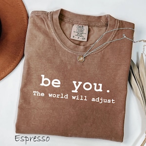 K&ouml;nnte beinhalten: Espressofarbenes T-Shirt mit dem wei&szlig;en Text "be you. The world will adjust". Das Shirt hat einen Rundhalsausschnitt und ein kleines Etikett am Ausschnitt. Eine silberne Halskette mit Anh&auml;nger liegt &uuml;ber dem Shirt.